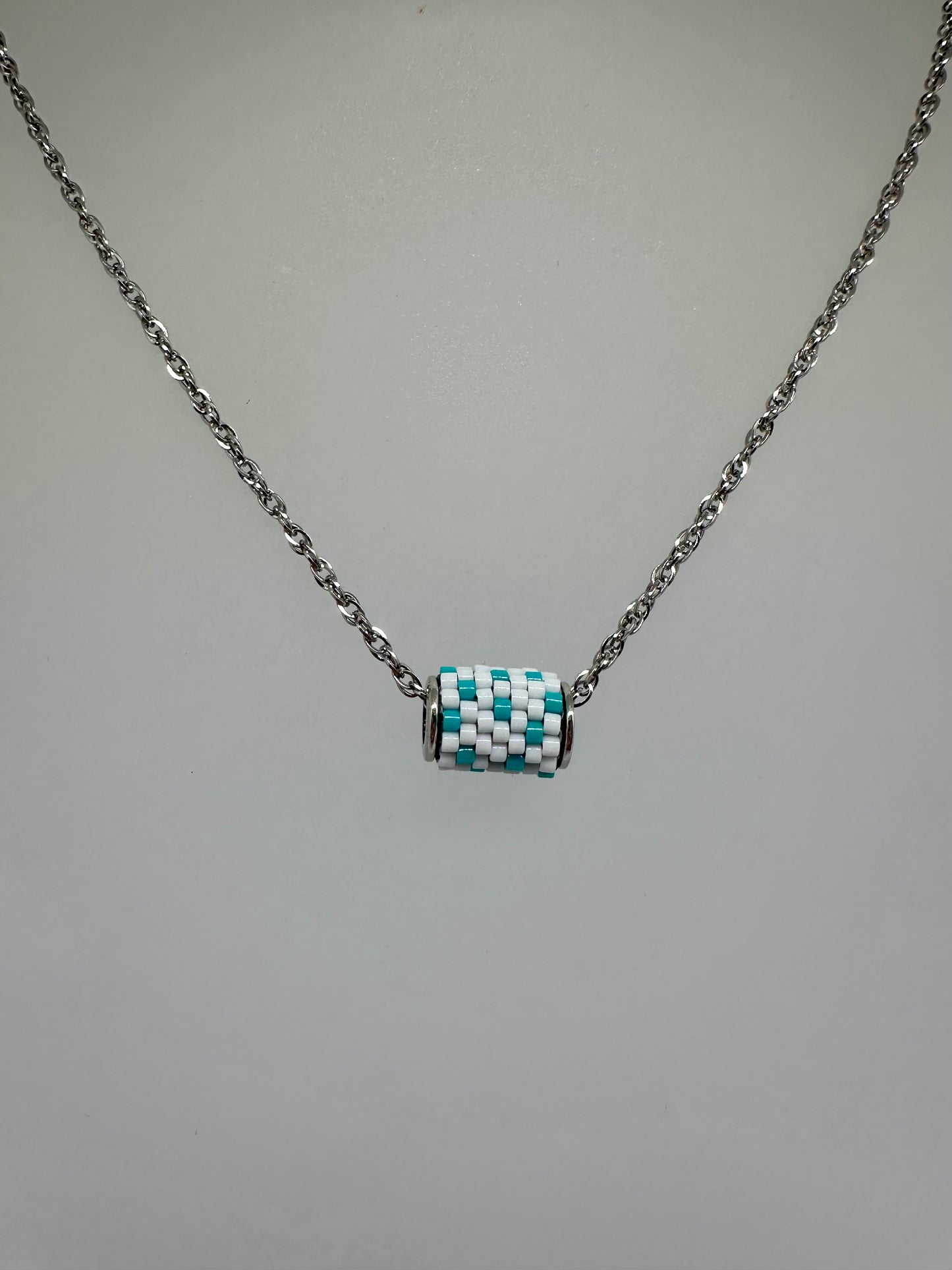 Close up of silver necklace with a beaded woven pendant with white and teal beads on a white bust 