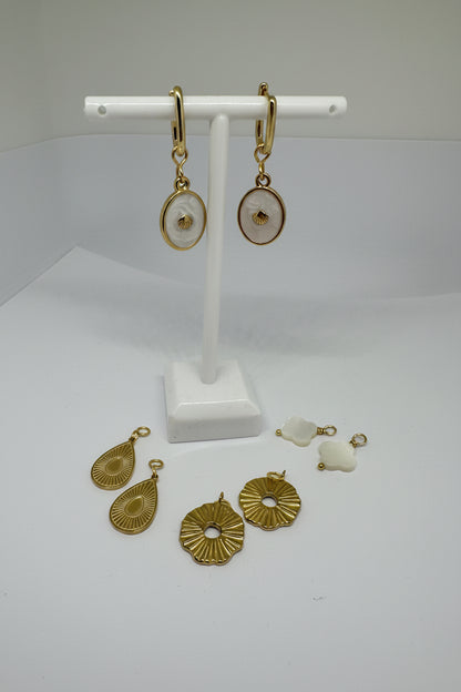 Gold huggie earrings with gold and white shell charms surrounded by interchangeable charms in gold and white on a white stand and surface.
