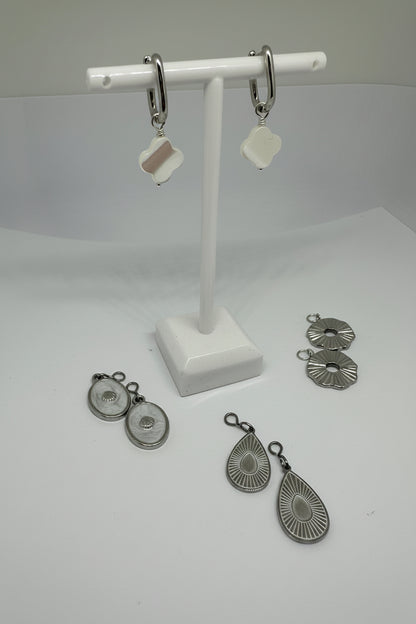 Silver huggie earrings with white mother of pearl clover charms surrounded by interchangeable charms in silver and white on a white stand and surface.