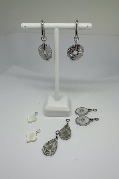 Silver huggie earrings with sunburst charms surrounded by interchangeable charms in silver and white on a white stand and surface.
